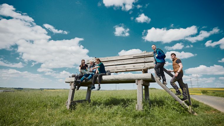 Wanderer sitzen auf einer XXL-Bank in Möntenich am Wanderweg Eifelhöhen und Brohlbachtal und genießen die Aussicht über die Eifellandschaft.