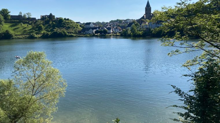 Ein ruhiger See mit klarem Wasser und grünen Bäumen im Vordergrund. Im Hintergrund sind Häuser und ein Kirchturm sichtbar.
