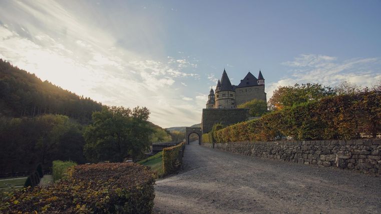 Eine malerische Landschaft mit einem Schloss im Hintergrund. Der Weg führt durch grüne Bäume und Sträucher in die Ferne.
