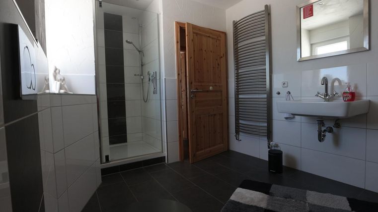 Ein Badezimmer mit einer Dusche und einem Holzregal. Die Wände sind hell gefliest und der Boden ist dunkler, ergänzt durch einen Teppich.