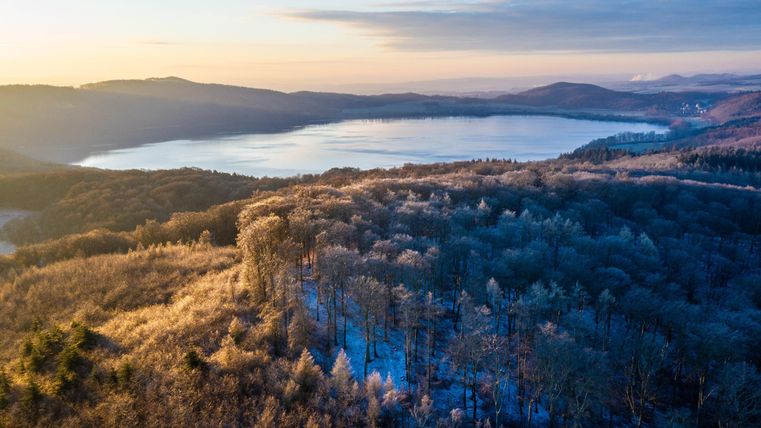 Eine malerische Winterlandschaft mit gefrorenen Bäumen und einem ruhigen See im Hintergrund. Die sanften Hügel rund um den See sind von frostigem Glanz bedeckt.