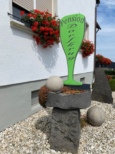 Een bord met de tekst "Pension Dorfkrug" staat voor een gebouw. Bloembakken en stenen versieren de ingang.