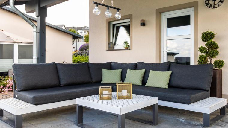 Eine moderne Gartenlounge mit einer großen, bequemen Couch und einem Tisch. Die Wohnung im Hintergrund und die Pflanzen sorgen für eine einladende Atmosphäre.