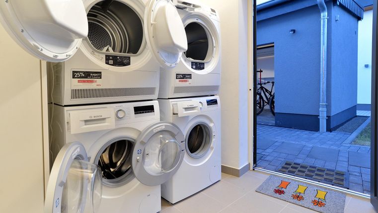 Eine moderne Waschküche mit einem Waschmaschine und einem Trockner übereinander. Die Tür führt nach draußen und ein Fußmatte liegt im Eingangsbereich.