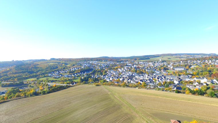 Eine großzügige Landschaft mit Feldern im Vordergrund und einer kleinen Stadt im Hintergrund. Die Umgebung ist von hügeliger Natur und buntem Herbstlaub geprägt.