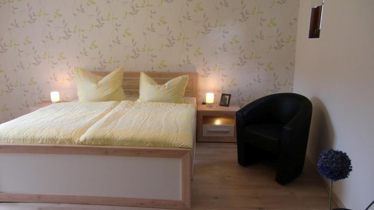 Une chambre avec un lit confortable et des couvertures jaunes. À côté du lit se trouve un fauteuil noir et un vase de fleurs.