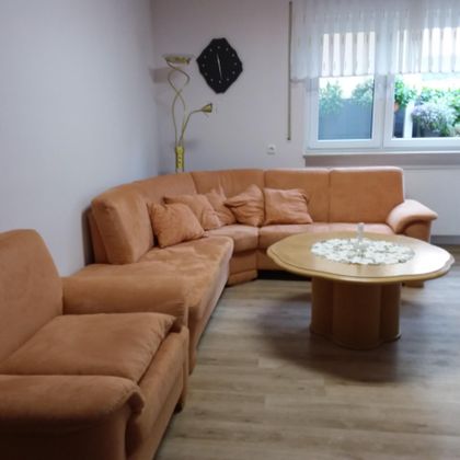 Eine gemütliche Sitzecke mit einem orangen Sofa und einem runden Tisch. Der Raum hat große Fenster, die für viel Licht sorgen.