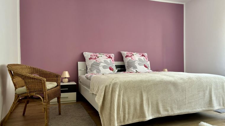 Ein gemütliches Schlafzimmer mit einem Doppelbett und bunten Kissen. Die Wand ist in sanftem Lila gestrichen, und ein Korbstuhl steht daneben.