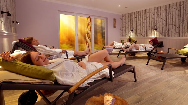 Ein ruhiger Wellnessbereich mit Liegen, in dem entspannte Menschen in Bademänteln liegen. Sanftes Licht und beruhigende Farben schaffen eine entspannende Atmosphäre.
