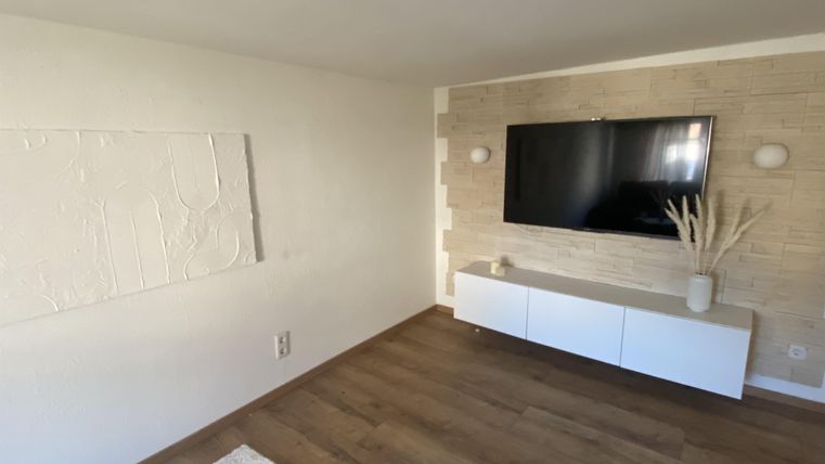 Ein modernes Wohnzimmer mit einem Flachbildfernseher und einer schlichten weißen Wohnwand. Die Wände sind in neutralen Farben gehalten, und der Boden ist aus Holz.