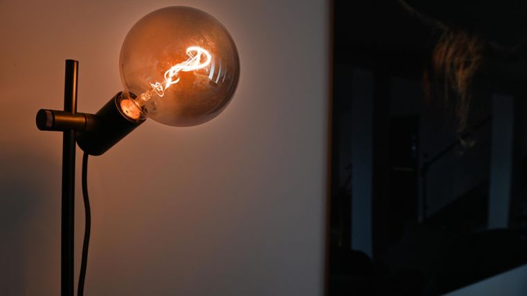 Eine moderne Stehlampe mit einer glühenden Glühbirne. Im Hintergrund ist eine verschwommene Innenraumansicht sichtbar.