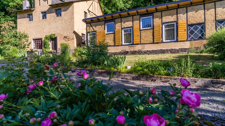 Un beau bâtiment caractéristique avec une annexe moderne. Au premier plan, des pivoines roses fleurissent dans un jardin soigné.