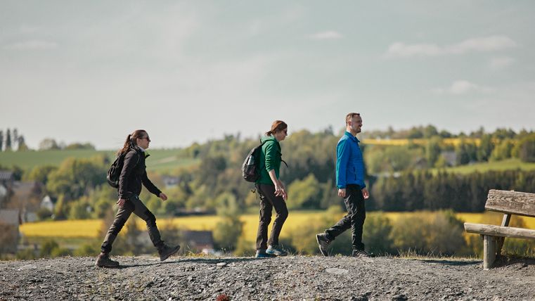Eine Gruppe von Wanderern die über dunkles Schiefergestein wandern. Im Hintergrund befindet sich eine Frühlingslandschaft.
