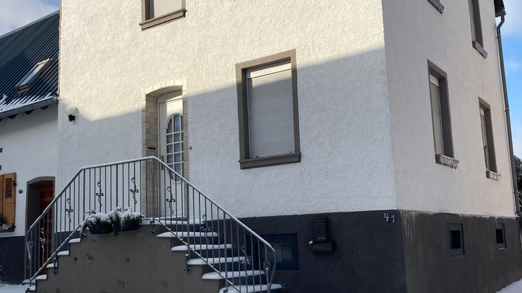 Ein weißes Haus mit einem schwarzen Sockel und einer Treppe. Um das Haus liegt Schnee, und der Himmel ist teilweise bewölkt.