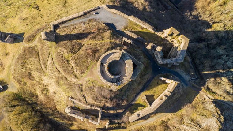 Eine Luftaufnahme einer alten Festungsruine mit kreisförmigen und geraden Mauern. Umgeben von grünen Hügeln und natürlicher Vegetation.
