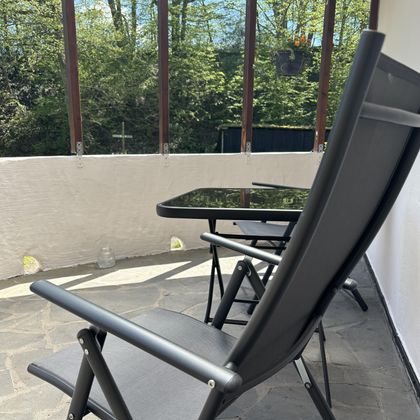 Ein entspannter Balkon mit einem klappbaren Stuhl und einem Tisch aus Glas. Im Hintergrund sind grüne Bäume zu sehen.