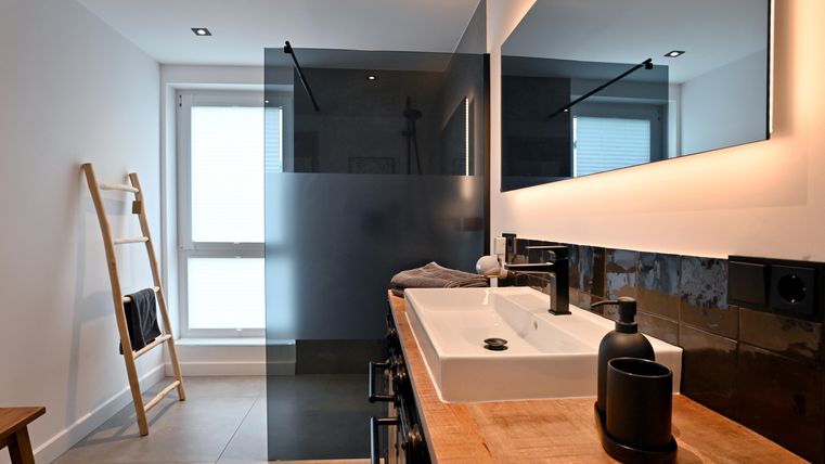 Ein modernes Badezimmer mit elegantem Waschbecken und dunklen Fliesen. Das Raumdesign ist minimalistisch und hell, mit einer großen Spiegelfläche.