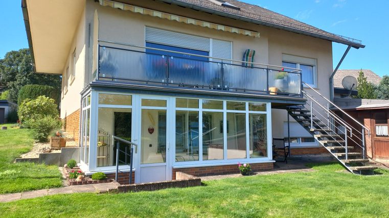 Ein modernes Haus mit einem großen Balkon und einer Treppe. Die Umgebung ist grün und gepflegt.