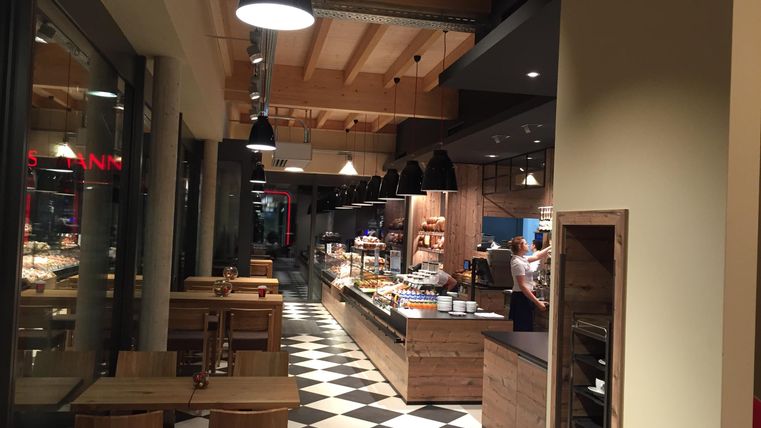 Ein gemütliches Café mit Holzinterieur und einer offenen Verkaufsfläche. Es sind Tische und Stühle vorhanden, sowie eine Auswahl an Speisen in der Vitrine.