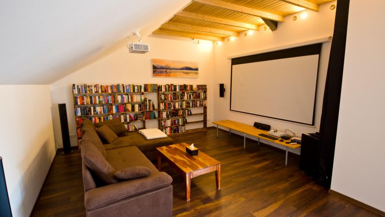 Ein gemütliches Wohnzimmer mit einem braunen Sofa und einem großen Bücherregal. An der Wand befindet sich ein Projektionsschirm und ein helles Holzdach.