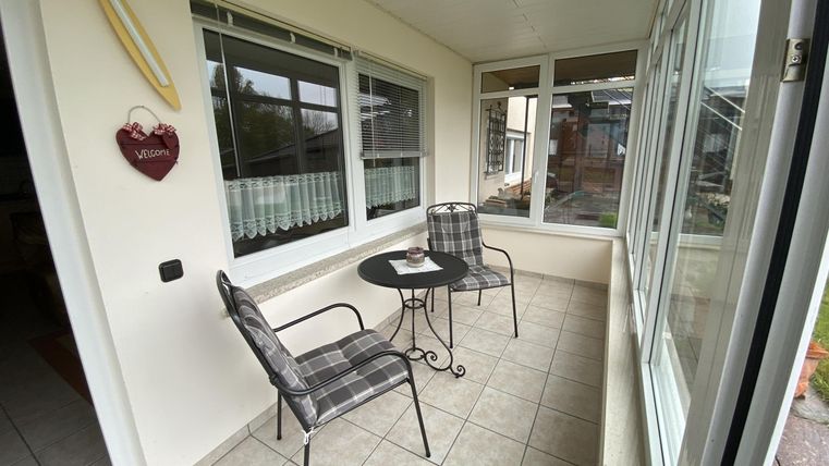Ein gemütlicher Balkon mit zwei Stühlen und einem kleinen Tisch. Die großen Fenster bieten viel Tageslicht und einen Blick nach draußen.