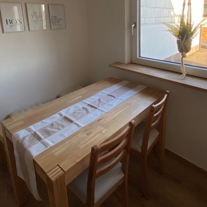 Ein einfacher Holztisch mit drei Stühlen steht in einem hellen Raum. Die Fenster bieten Tageslicht und eine kleine Pflanze hängt dekorativ auf der Fensterbank.