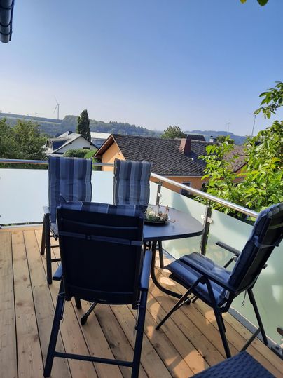 Ein gemütlicher Balkon mit einem Tisch und Stühlen. Im Hintergrund sind Häuser und ein klarer Himmel zu sehen.