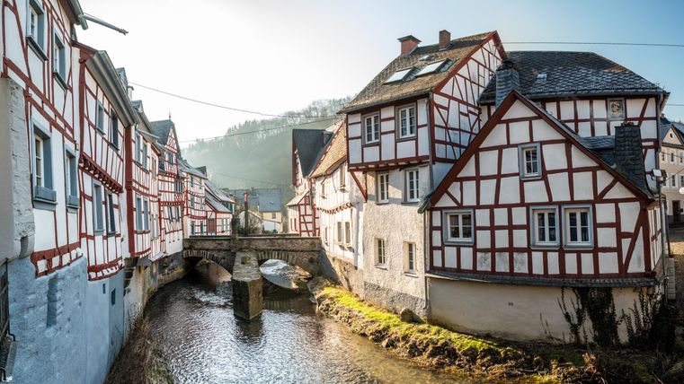 Eine malerische Stadt mit Fachwerkhäusern und einem kleinen Fluss. Im Hintergrund sind sanfte Hügel und blauer Himmel zu sehen.