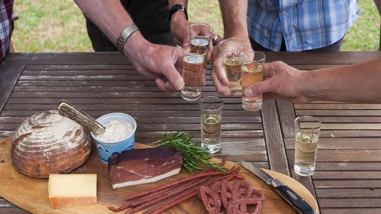 Un groupe de personnes lève des verres à shot pour trinquer. Sur la table, il y a une planche rustique avec du pain, du fromage, de la charcuterie et des herbes fraîches.