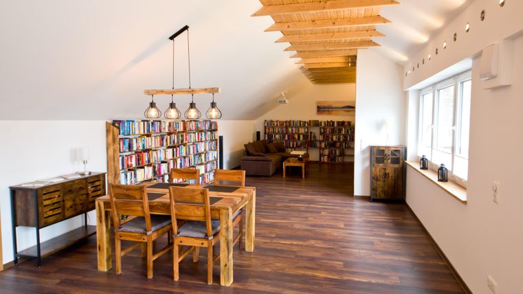 Ein moderner Raum mit Holzböden und einem großen Bücherregal. Ein Essbereich mit einem Tisch und Stühlen ist gut beleuchtet und lädt zum Lesen ein.