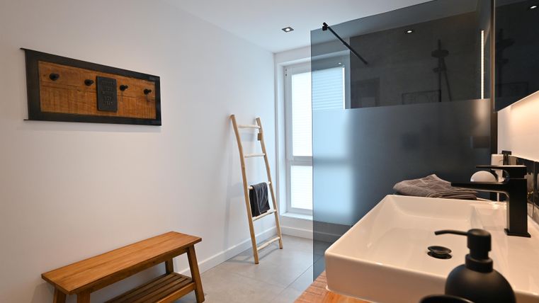 Ein modernes Badezimmer mit klaren Linien und neutralen Farben. Es verfügt über ein Waschbecken, eine gläserne Duschwand und eine Holzbank.