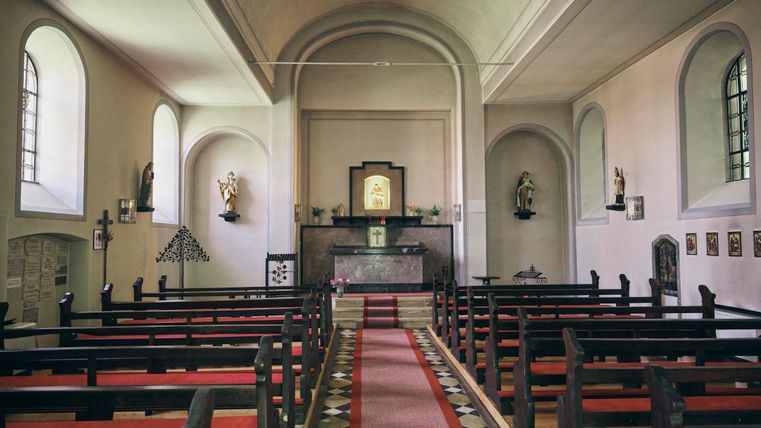 Eine ruhige Kirche mit Holzbänken und einem klaren Gang zum Altar. An den Wänden sind religiöse Statuen und Bilder zu sehen.