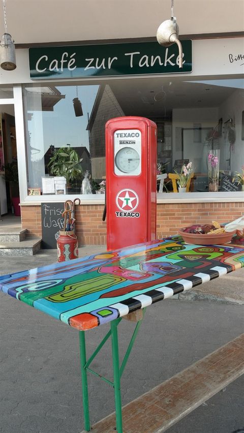 Ein bunt bemalter Tisch steht vor einem Café mit der Aufschrift „Café zur Tanke“. Im Hintergrund ist eine alte Texaco-Benzinpumpe zu sehen.