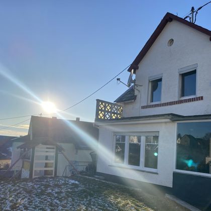 Ein helles Haus mit großen Fenstern, das im Sonnenlicht strahlt. Im Hintergrund ist ein klarer Himmel zu sehen.