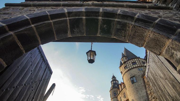 Ein historisches Tor mit einer Laterne, das auf eine beeindruckende Burg blickt. Der Himmel ist klar und blau, was eine idyllische Atmosphäre schafft.