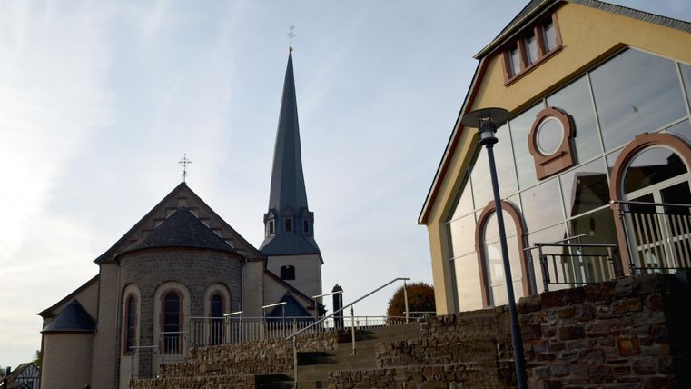 Eine Kirche mit einem hohen Turm steht neben einem modernen Gebäude. Die Landschaft ist ruhig und einladend.