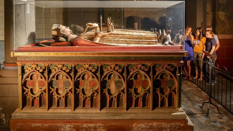 Ein historisches Grabmal in einem Museum, umgeben von einer Glasvitrine. Besucher stehen in der Nähe und betrachten die Ausstellungsstücke.