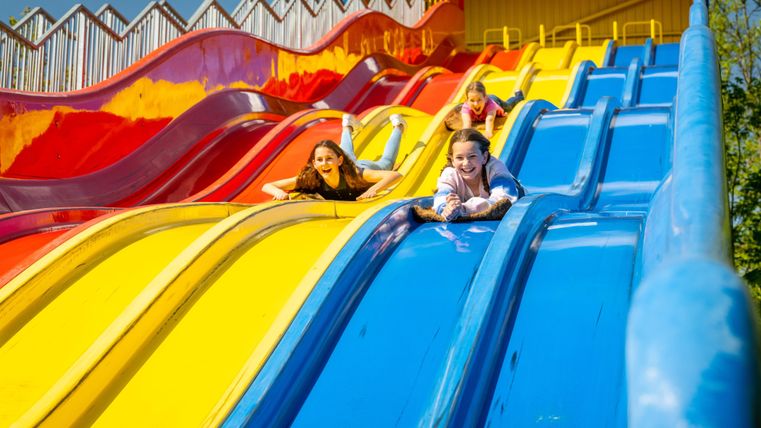 Drei Kinder rutschen fröhlich die bunten Rutschen hinunter. Der sonnige Himmel und die lebhaften Farben machen die Szene einladend und verspielt.