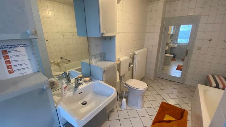 Ein Badezimmer mit weißen Fliesen und einem großen Spiegel. Es enthält eine Badewanne, ein Waschbecken und orangefarbene Teppiche.