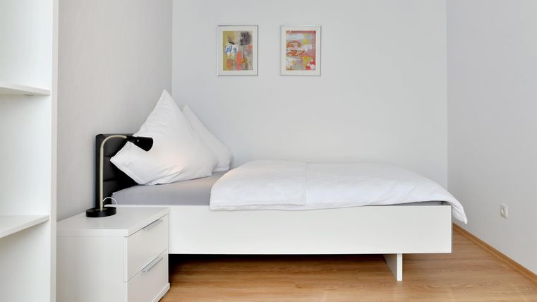 Ein helles Schlafzimmer mit einem Einzelbett und einem Nachttisch. An der Wand hängen zwei Bilder, und der Boden ist aus Holz.