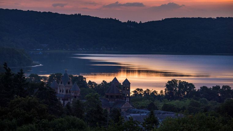 Ein malerischer Sonnenuntergang über einem ruhigen See, umgeben von sanften Hügeln. Im Vordergrund ist ein historisches Gebäude sichtbar, das eine geheimnisvolle Atmosphäre erzeugt.