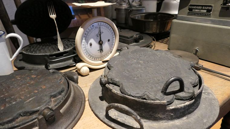 Eine Sammlung von alten Küchenutensilien, darunter ein Waffeleisen, eine Küchenwaage und verschiedene Gusseisenformen. Die Gegenstände stehen auf einem Holztisch und vermitteln ein nostalgisches Gefühl.