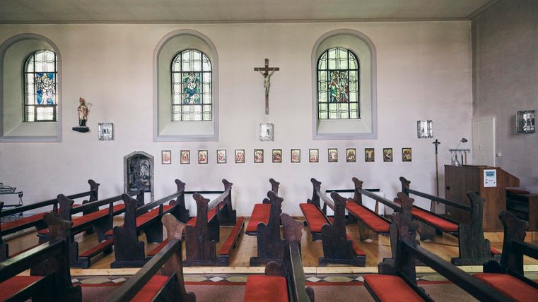 Eine ruhige Kirche mit Holzbankreihen und roten Polstern. An den Wänden hängen Bilder und es gibt bunte Fenster, die Licht hereinlassen.