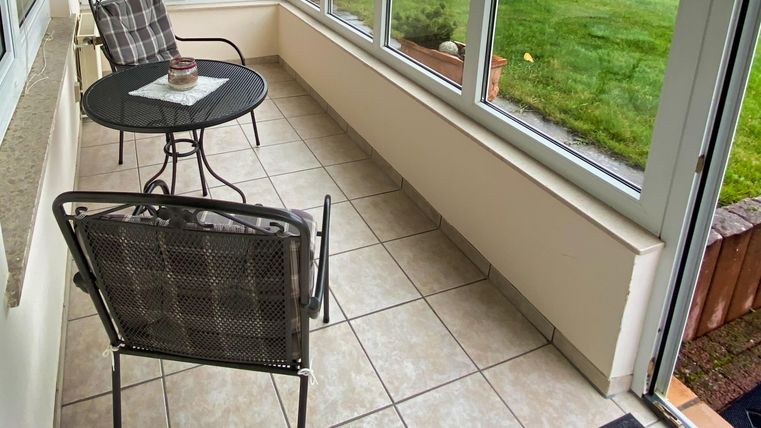 Ein heller Wintergarten mit Fliesenboden und zwei Stühlen. Am Tisch steht ein kleiner Dekorationsgegenstand, im Hintergrund ist der Garten sichtbar.