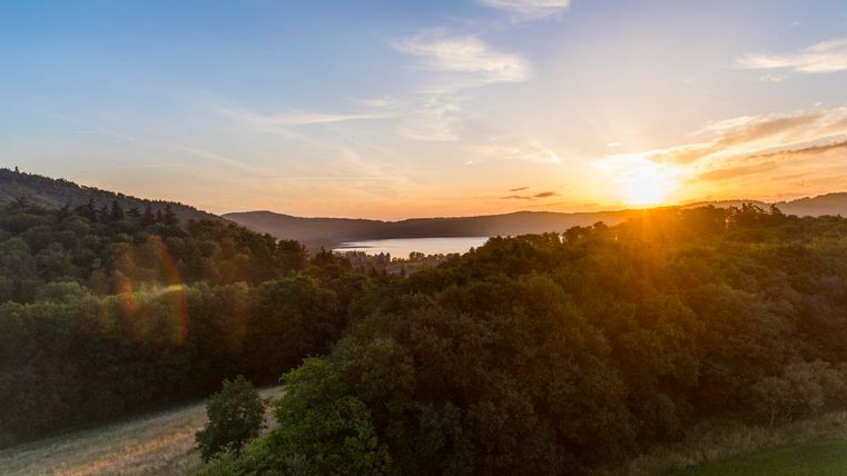 Ein malerischer Sonnenuntergang über einer hügeligen Landschaft. Grüne Wälder und Felder umgeben einen ruhigen See.
