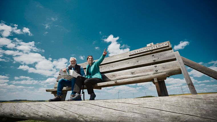 Ein großes Holzbank in der Landschaft mit zwei Personen, die fröhlich sitzen und in die Kamera zeigen. Der Himmel ist blau mit wenigen weißen Wolken.