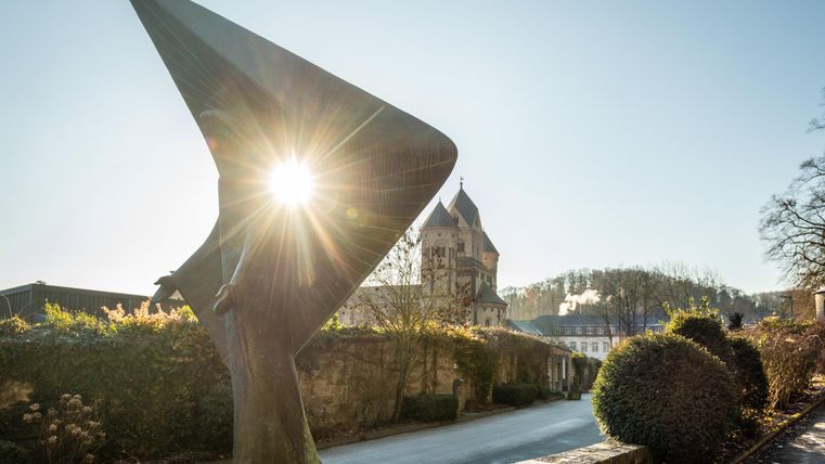 Eine moderne Statue mit spitzen Formen, die von der Sonne beleuchtet wird. Im Hintergrund ist ein historisches Gebäude und eine ruhige Straße zu sehen.