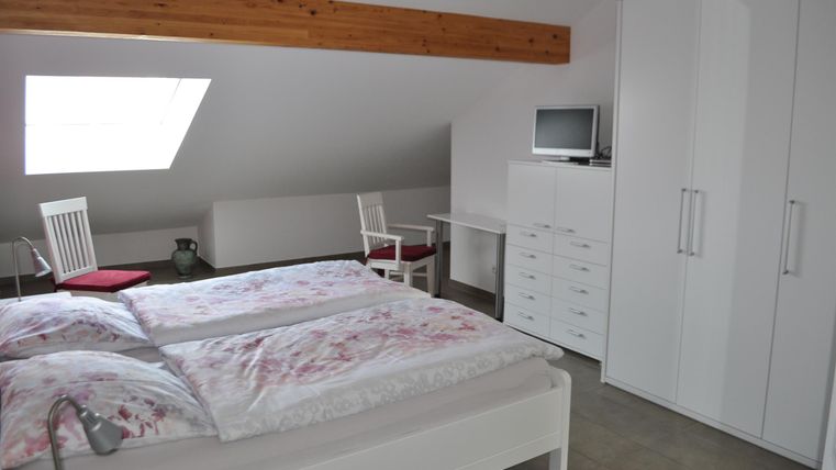 Ein helles Schlafzimmer mit einem Doppelbett und einem großen Schrank. Es gibt einen Tisch und einen Stuhl sowie einen Fernseher an der Wand.