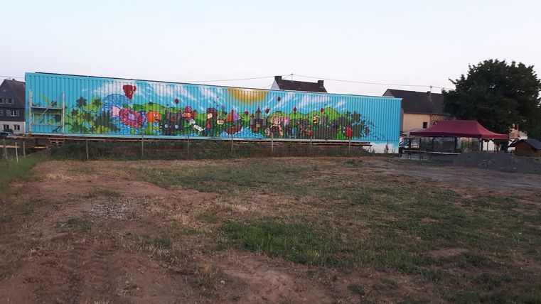 Eine bunte Wandmalerei mit Blumen und sonnigen Motiven. Im Vordergrund sind grüne Wiesen und ein paar Bäume sichtbar.