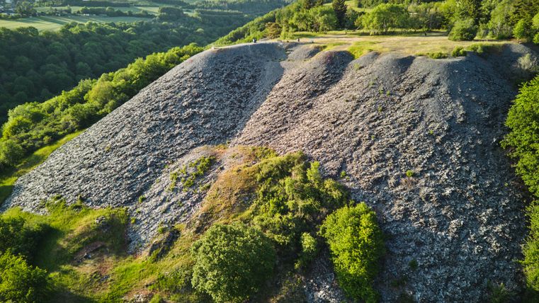 Aussichtspunkt Maria Schacht in der Eifel: Im Vordergrund ein großer, grauer Schieferhang mit spärlichem Bewuchs, umgeben von grünen Bäumen. Dahinter eine ebene Wiese mit Bäumen, im Hintergrund bewaldete Hügel und Felder unter klarem Himmel.